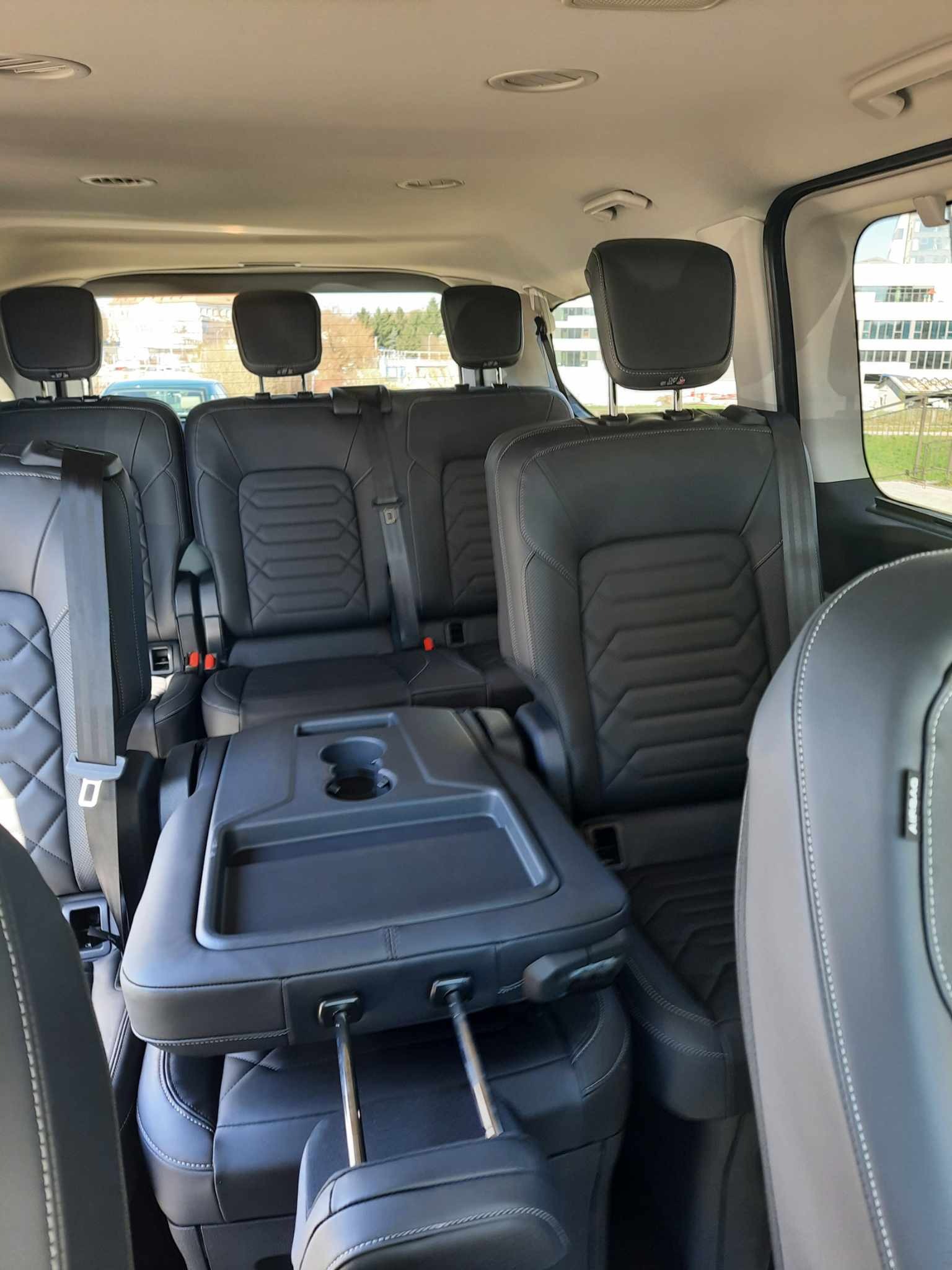 Wnętrze przestronnego vana z trzema rzędami czarnych skórzanych foteli, rozkładany stolik z uchwytem na napoje, widok z perspektywy pasażera.