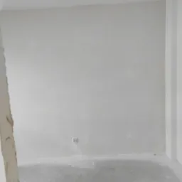 Przygotowane do malowania wnętrze z gładzią na ścianach i betonową wylewką na podłodze, widoczne gniazdko elektryczne i narożnik ściany w prawym górnym rogu.