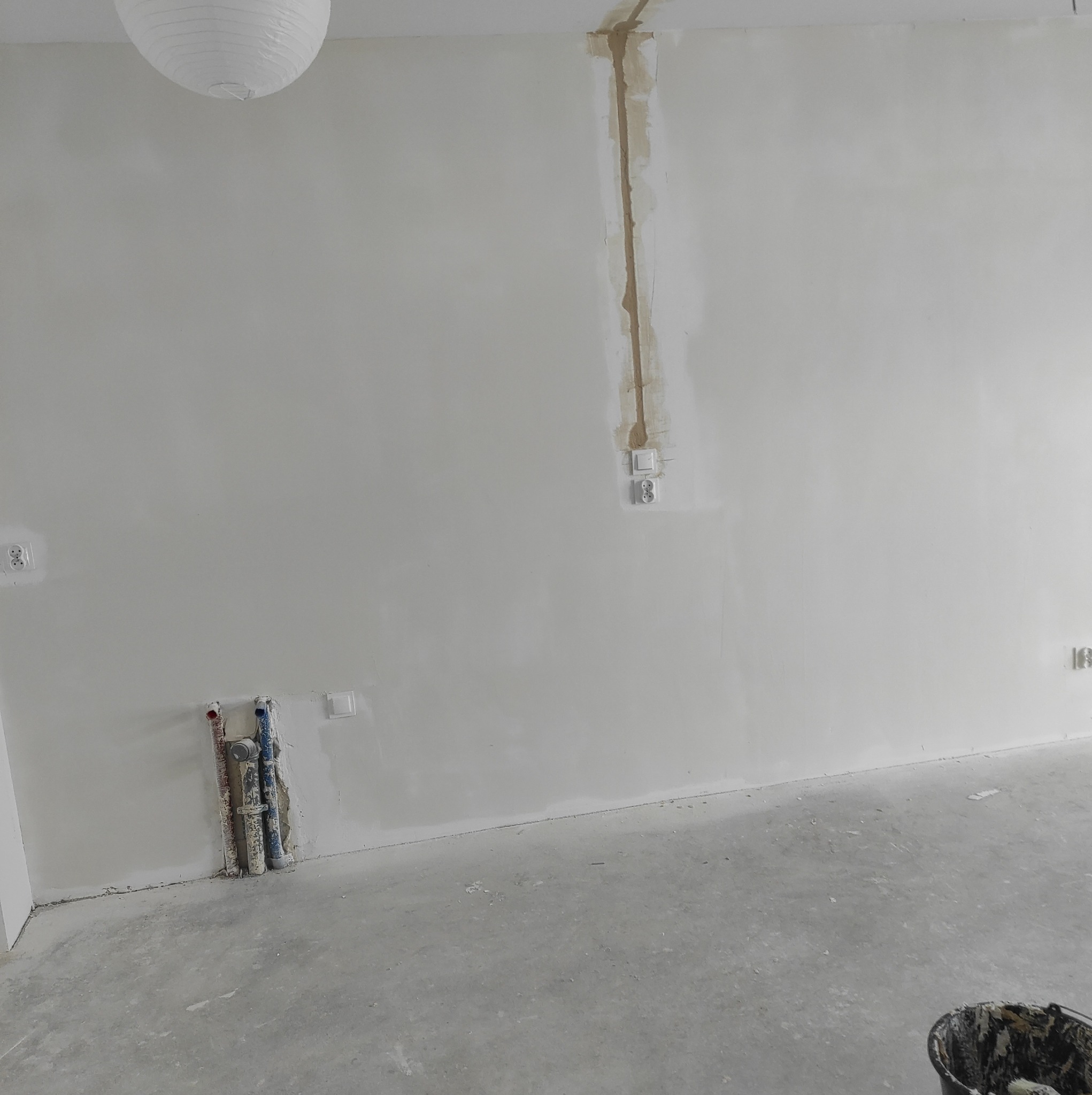 Wnętrze w trakcie prac przygotowawczych do malowania, widoczne ślady szpachlowania przy instalacji elektrycznej i rurach, surowy beton na podłodze, biała lampa papierowa pod sufitem.