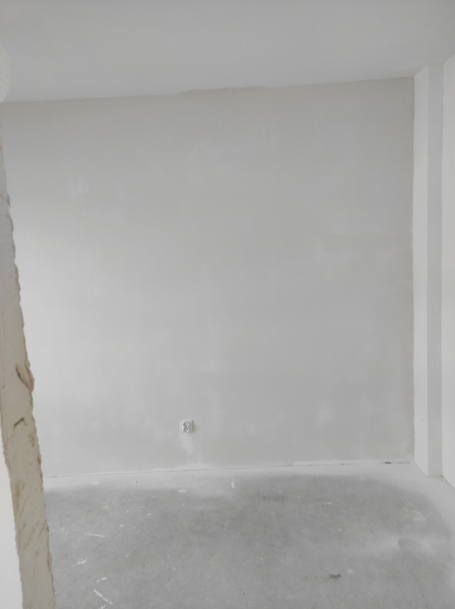 Przygotowane do malowania wnętrze z gładzią na ścianach i betonową wylewką na podłodze, widoczne gniazdko elektryczne i narożnik ściany w prawym górnym rogu.