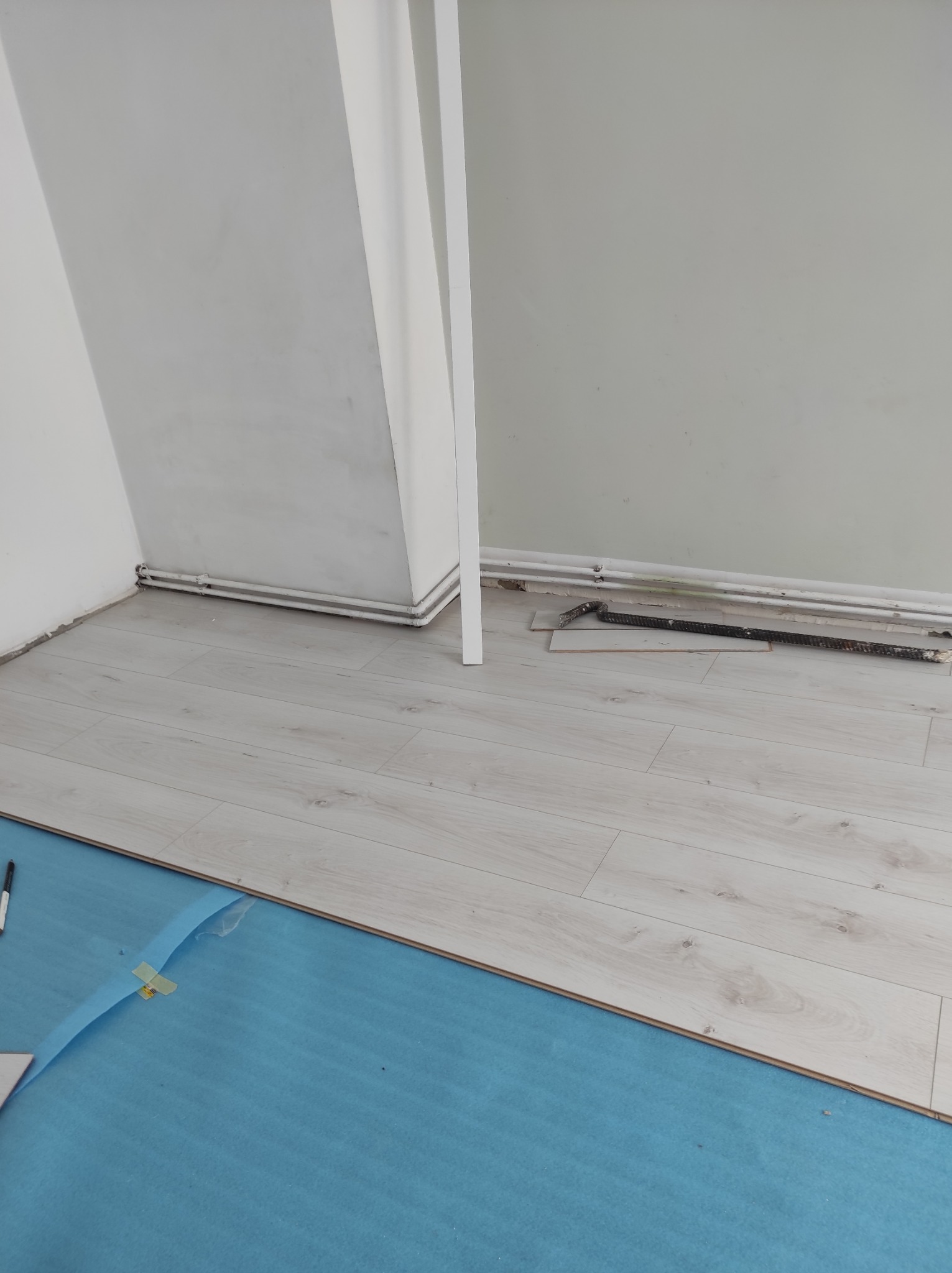 Układanie jasnych paneli podłogowych na niebieskiej piance izolacyjnej, widoczne przycięte panele i narzędzia przy ścianie.