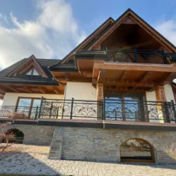 Dom w stylu góralskim z balkonami ozdobionymi kutymi, czarnymi balustradami z motywem roślinnym, widok z dołu na tle błękitnego nieba z chmurami.