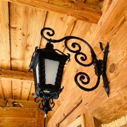 Metalowe Podhale Krzysztof Łowisz - Kuta, czarna lampa zewnętrzna z ozdobnymi spiralami, zamontowana na drewnianej ścianie domu z bali.