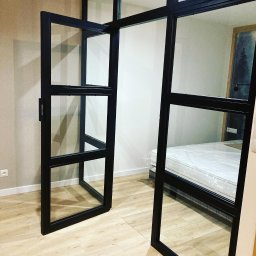 Metalowe Podhale Krzysztof Łowisz - Szklana ścianka działowa z czarnymi profilami, częściowo otwarta, oddzielająca sypialnię z łóżkiem w folii ochronnej, widoczny parkiet i gniazdko elektryczne na ścianie.