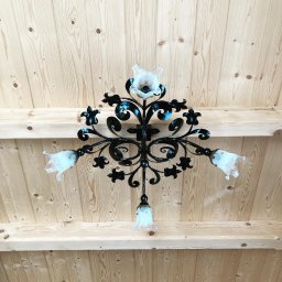 Metalowe Podhale Krzysztof Łowisz - Kuta, metalowa konstrukcja z motywami roślinnymi i szklanymi, kwiatowymi kloszami, zamontowana na jasnym, drewnianym suficie z widocznymi belkami.