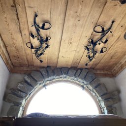 Metalowe Podhale Krzysztof Łowisz - Drewniany sufit z dwoma kutymi, metalowymi dekoracjami w kształcie roślinnym nad łukowym oknem z kamienną ramą, widok z dołu.