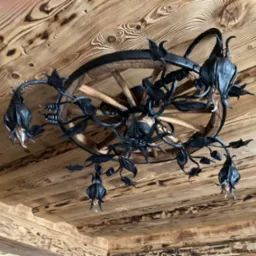 Ozdobny żyrandol z kutego metalu w kształcie kwiatów, zawieszony na drewnianym kole od wozu, zamontowany na drewnianym suficie z widocznymi słojami.