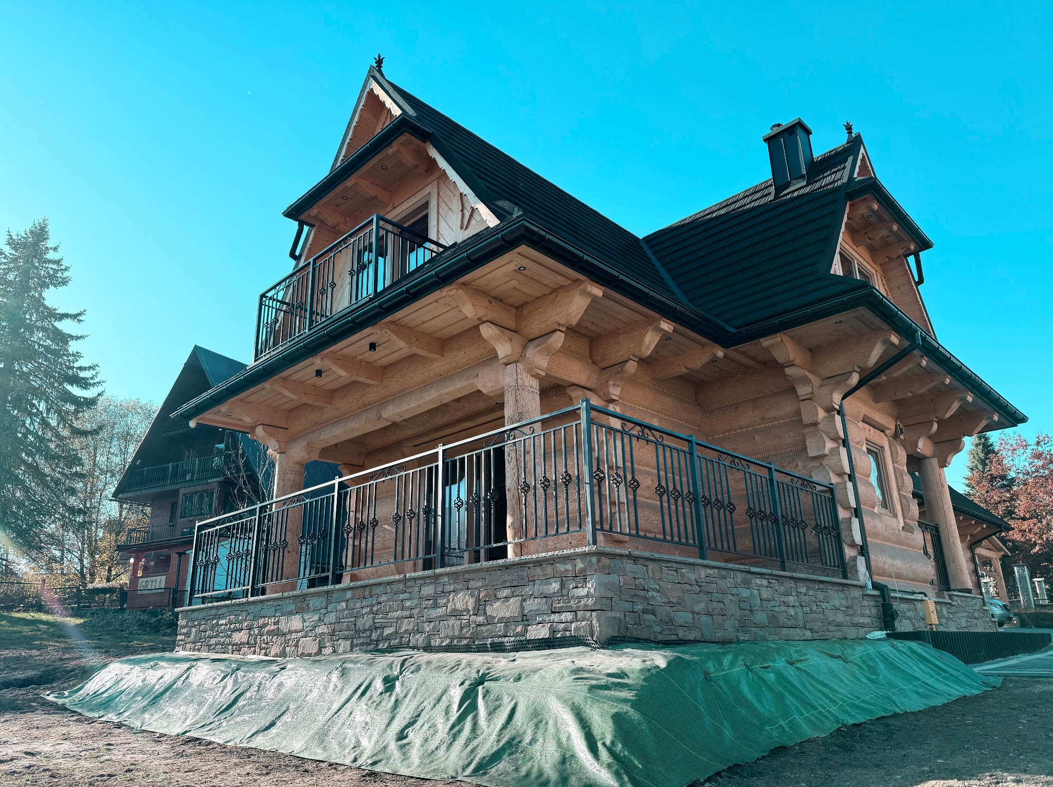 Dwukondygnacyjny dom z bali w stylu góralskim, z czarną dachówką i metalowymi balustradami balkonowymi z ozdobnymi elementami, posadowiony na kamiennej podmurówce, otoczony zieloną tkaniną ochronną.