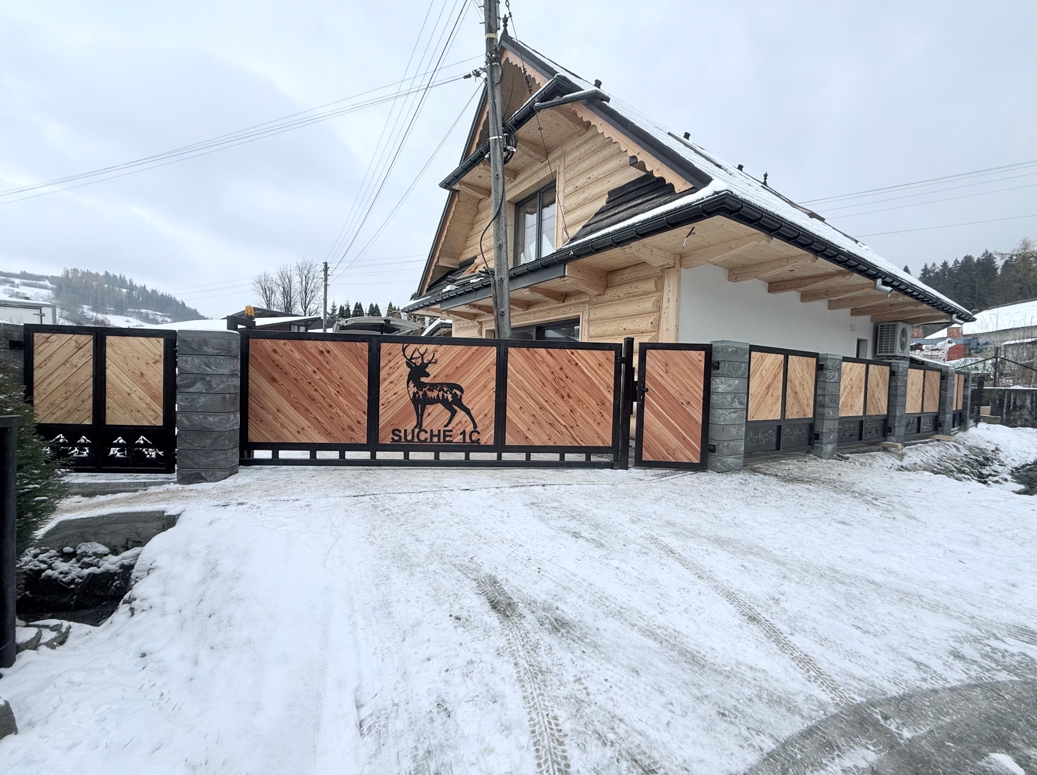 Nowoczesna brama wjazdowa z drewnianymi panelami ułożonymi w jodełkę, z motywem jelenia wyciętym laserowo, prowadząca do drewnianego domu w stylu góralskim, zimowy krajobraz.