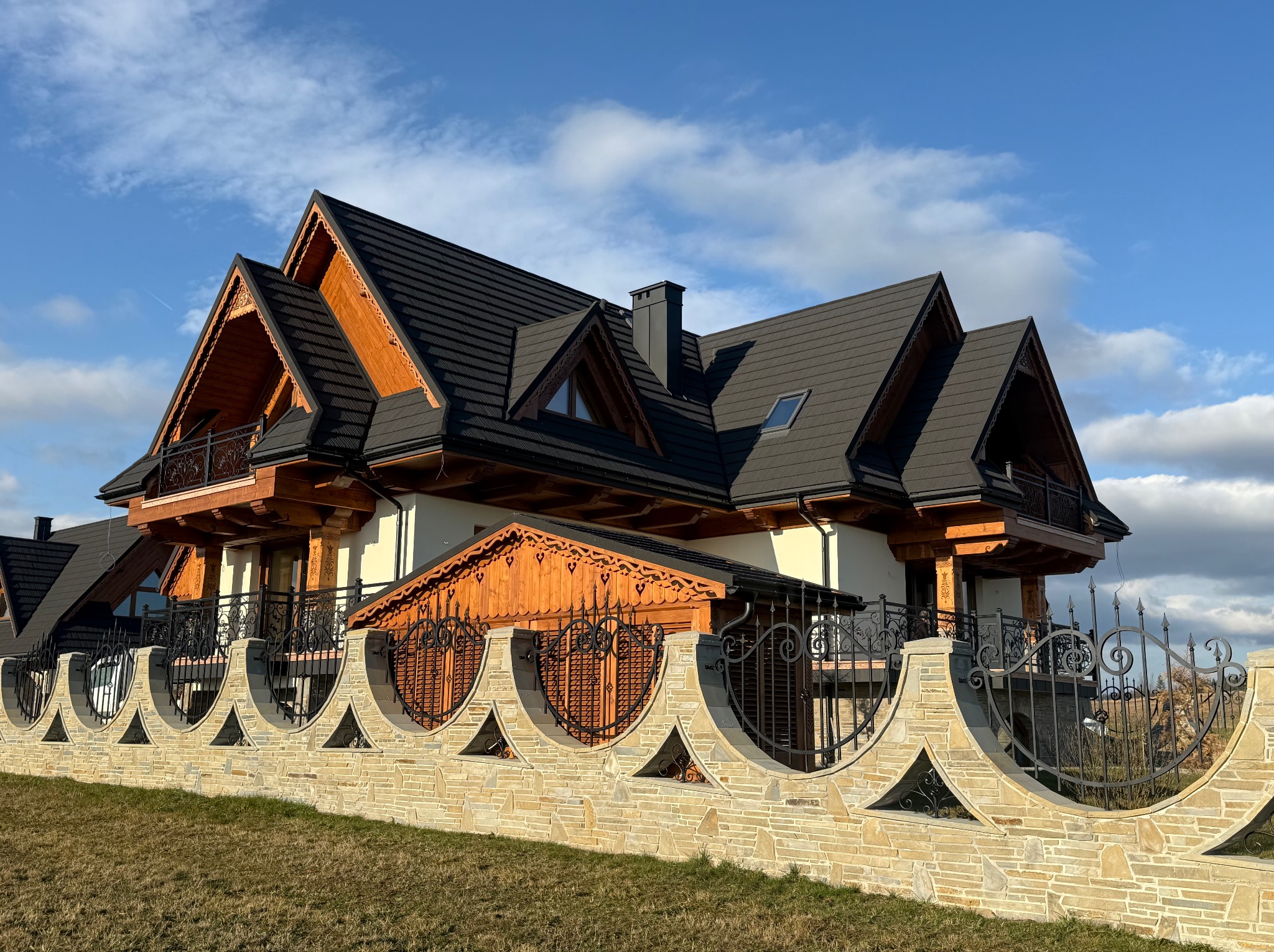 Nowoczesny dom z bali z czarnym dachem krytym dachówką, ozdobnymi drewnianymi wykończeniami i kamienno-metalowym ogrodzeniem z dekoracyjnymi elementami w kształcie serc i trójkątów.