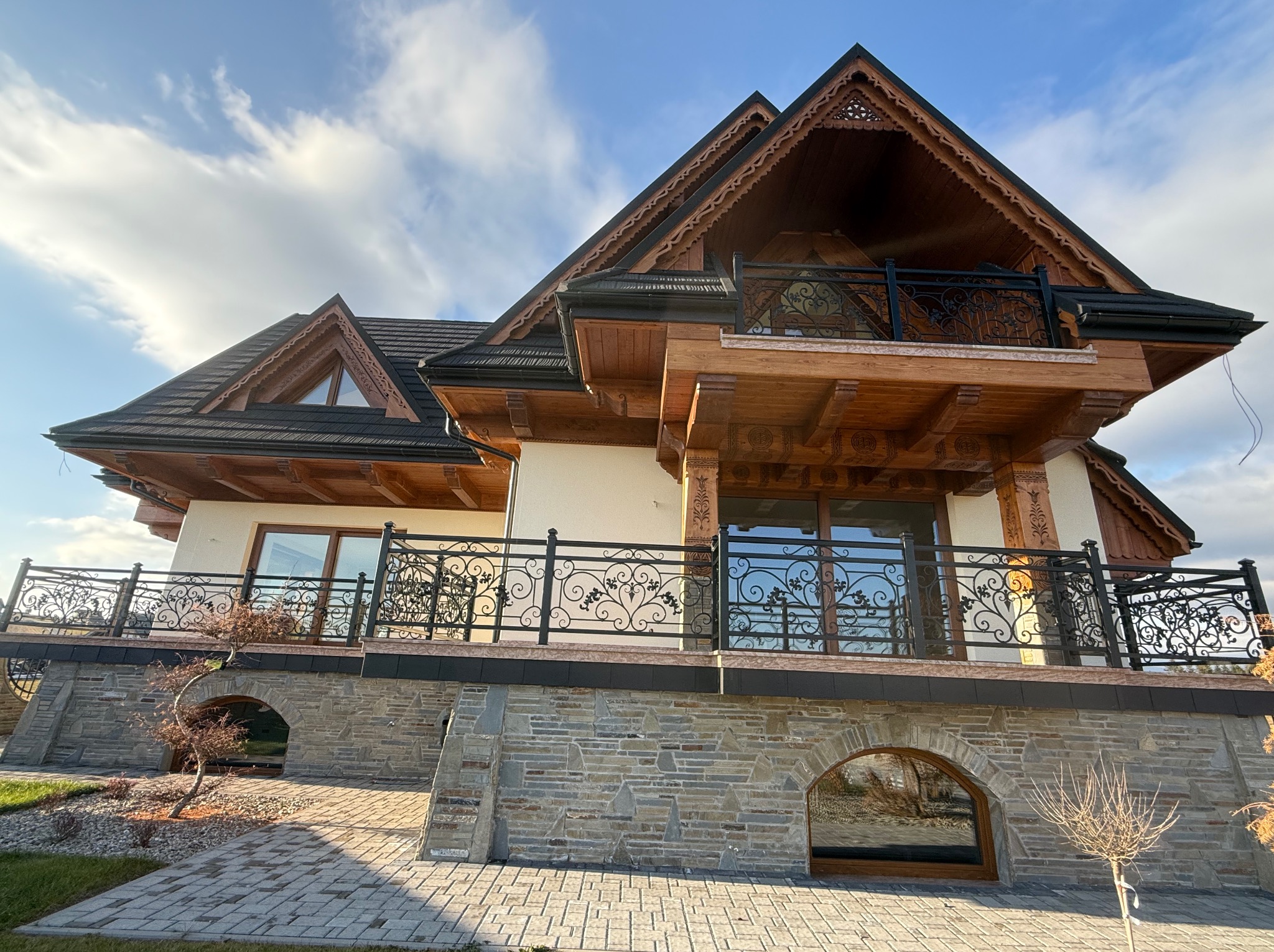 Dom w stylu góralskim z balkonami ozdobionymi kutymi, czarnymi balustradami z motywem roślinnym, widok z dołu na tle błękitnego nieba z chmurami.