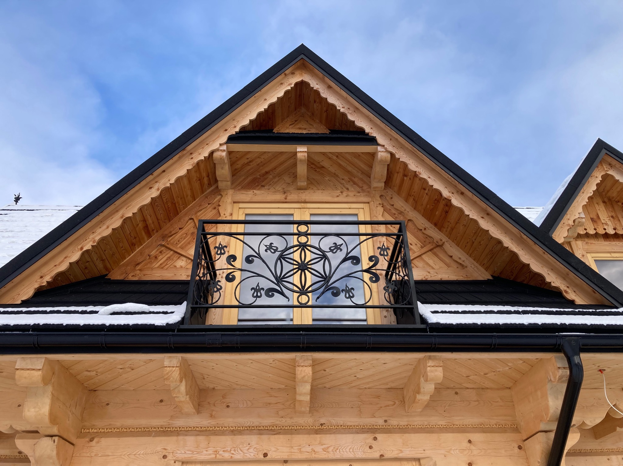 Drewniany balkon z czarną, ozdobną metalową balustradą na tle błękitnego nieba, widok z dołu na szczyt budynku.