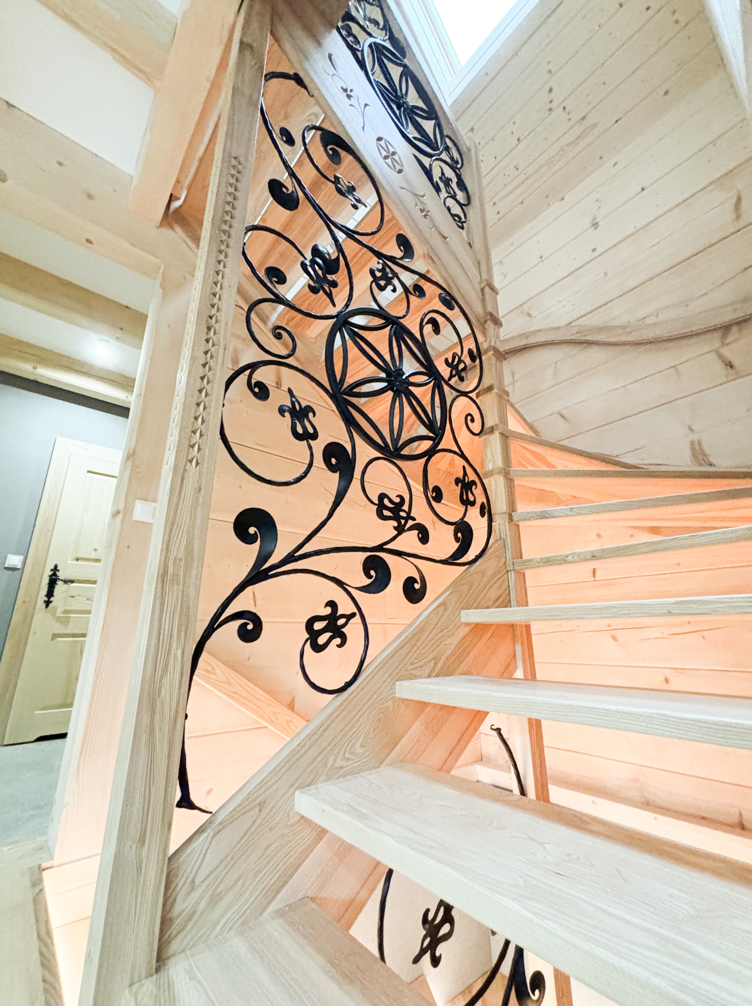 Drewniane schody spiralne z dekoracyjną, kutą, czarną metalową konstrukcją zabezpieczającą i oświetleniem LED pod stopniami, w domu wykończonym boazerią.