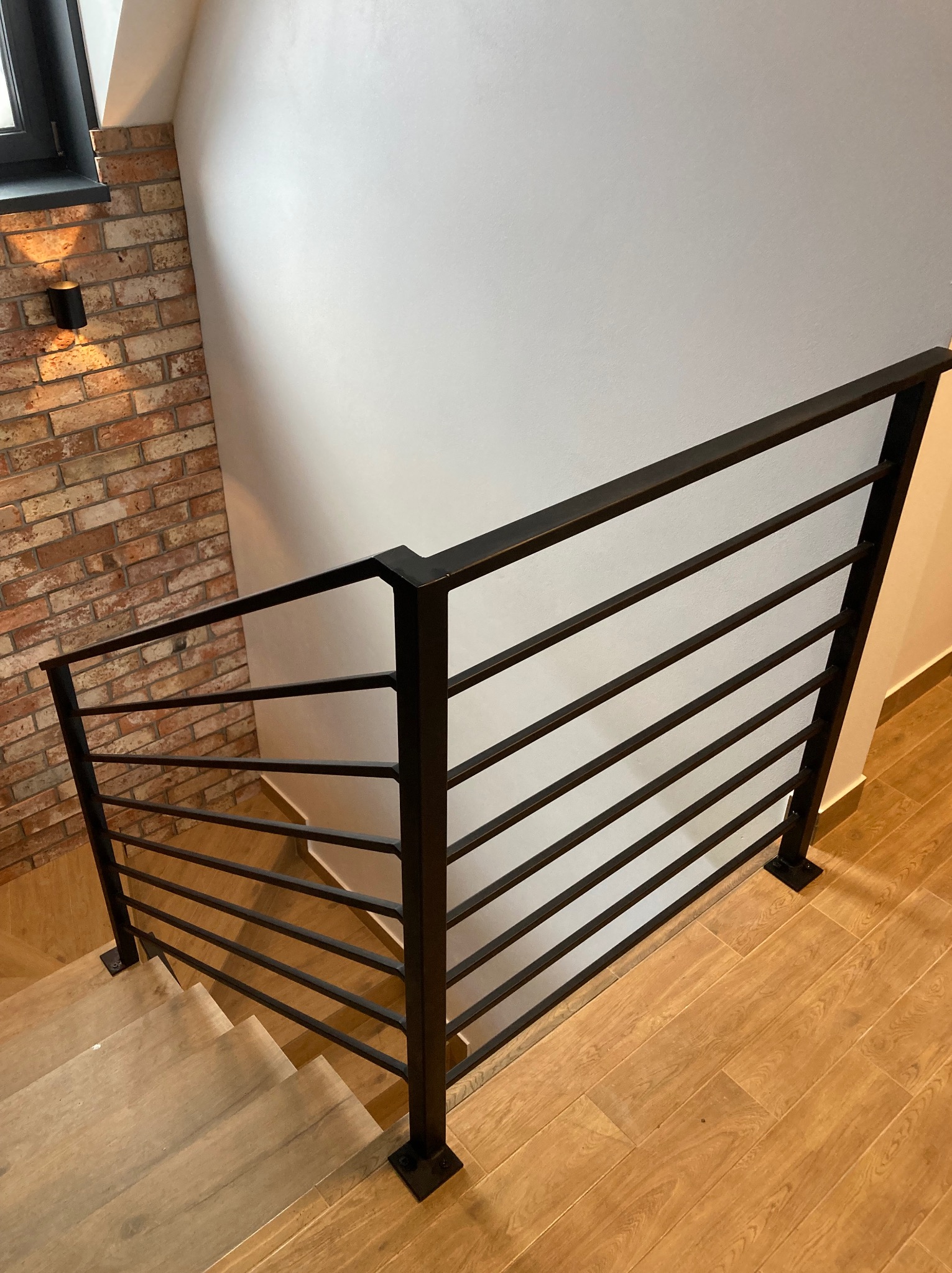 Nowoczesna, czarna, metalowa balustrada schodowa z poziomymi prętami, zamontowana wewnątrz budynku, widoczna na tle ściany z cegły i białej ściany, z drewnopodobnymi płytkami na podłodze.