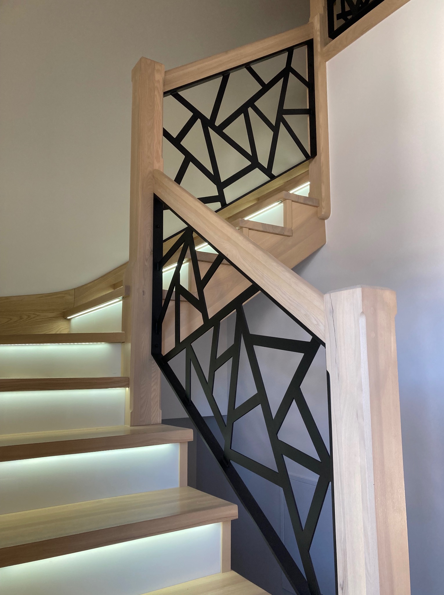 Nowoczesne schody drewniane z podświetleniem LED stopni i czarną, geometryczną konstrukcją zabezpieczającą, widok z dołu.