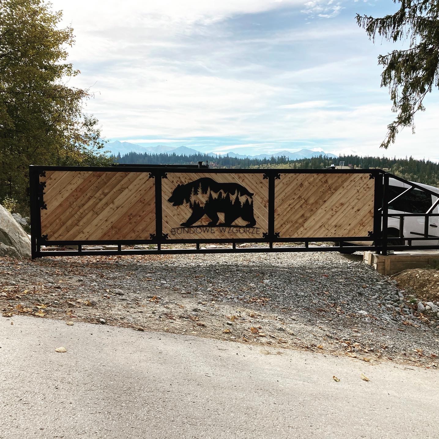 Drewniana brama wjazdowa z metalową ramą i dekoracyjnym motywem niedźwiedzia z drzewami, widok na góry w tle, styl rustykalny.