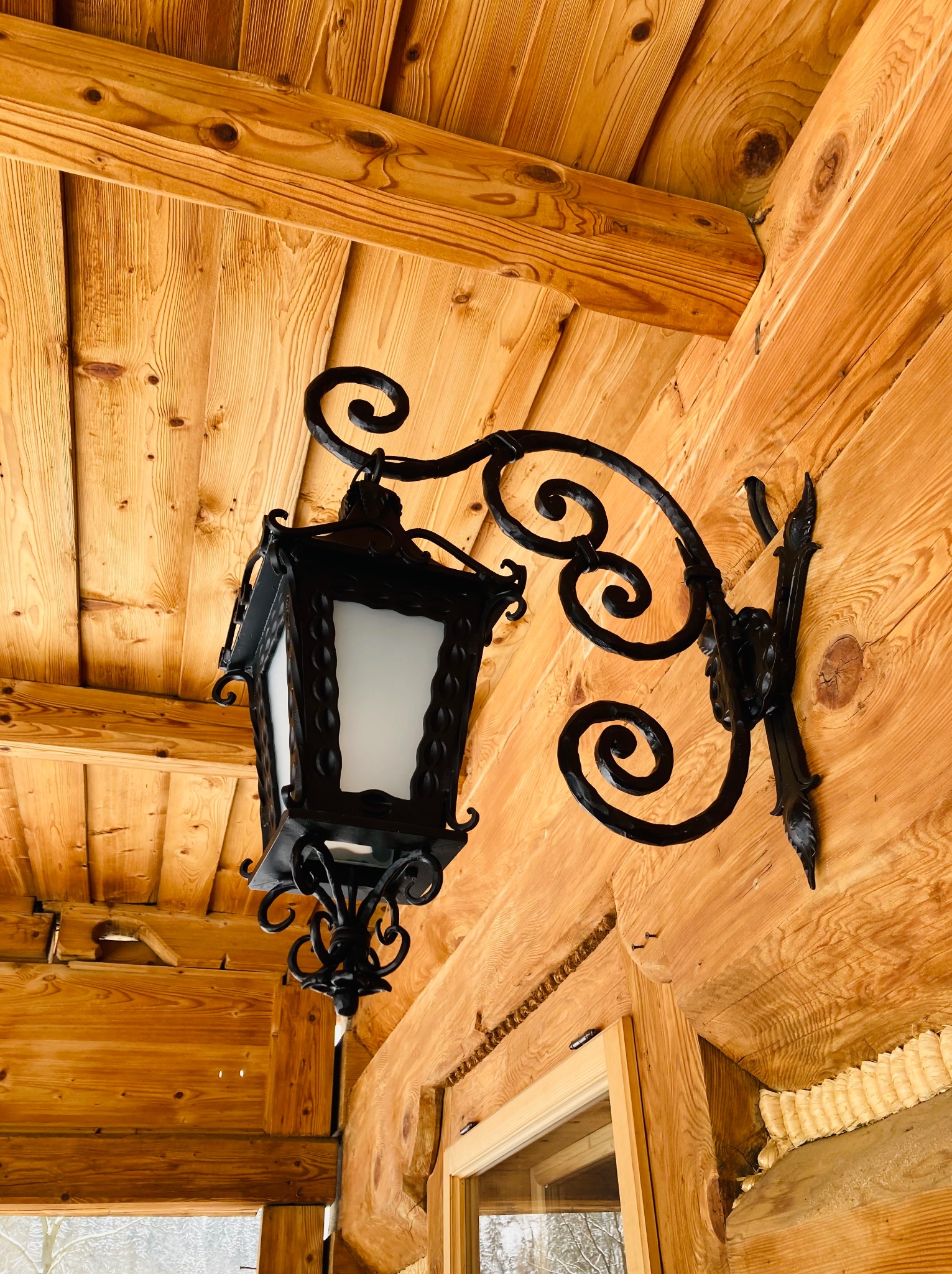Kuta, czarna lampa zewnętrzna z ozdobnymi spiralami, zamontowana na drewnianej ścianie domu z bali.