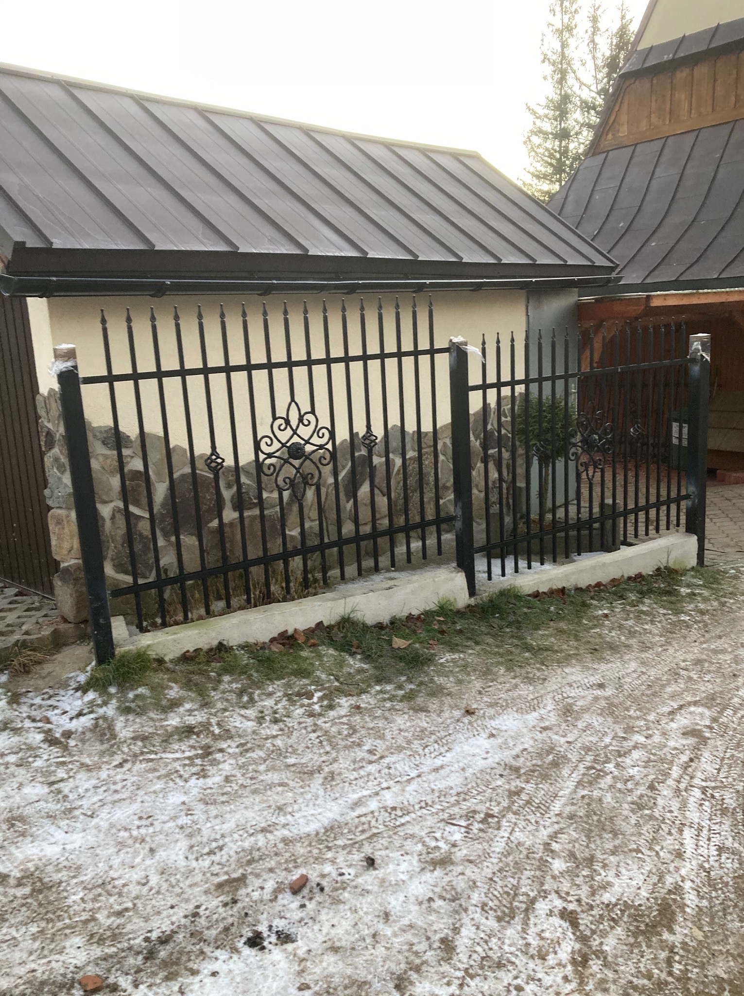 Czarne metalowe ogrodzenie z ozdobnymi elementami na podmurówce z kamienia, w tle budynek z ciemnym dachem, częściowo pokryty śniegiem teren.
