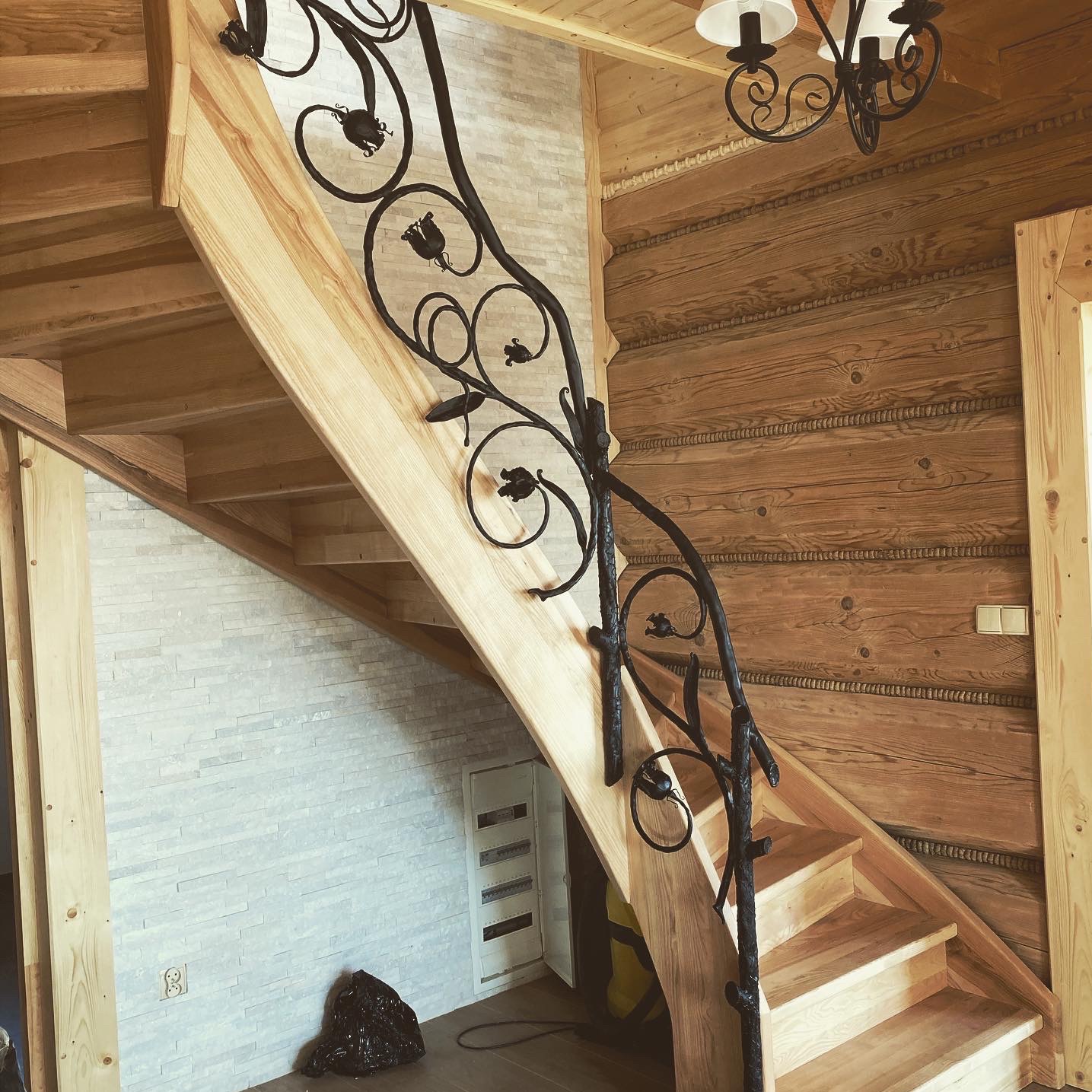 Drewniane schody z kutą, ozdobną balustradą w kształcie stylizowanych kwiatów, w rustykalnym wnętrzu z boazerią i kamienną ścianą.