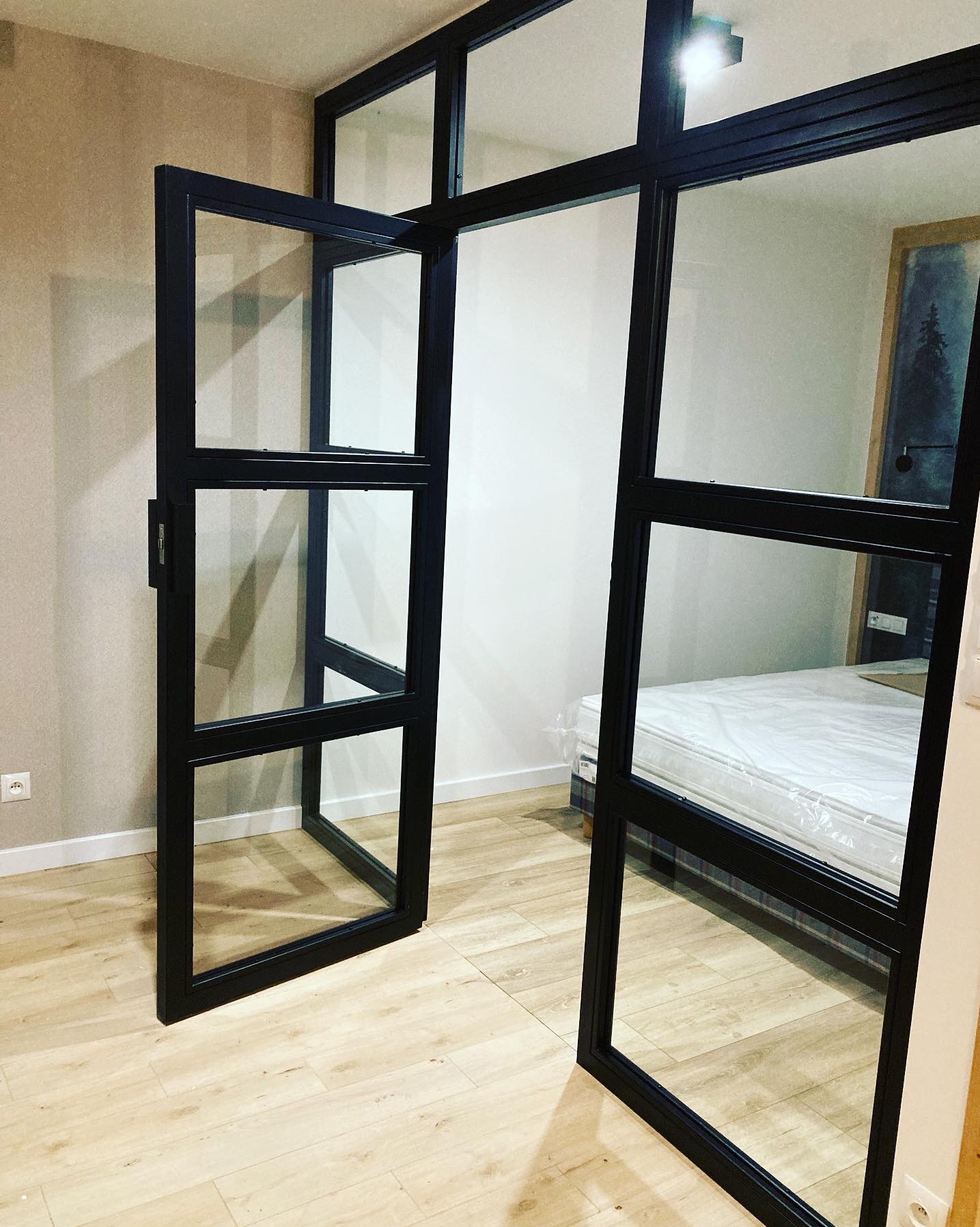 Szklana ścianka działowa z czarnymi profilami, częściowo otwarta, oddzielająca sypialnię z łóżkiem w folii ochronnej, widoczny parkiet i gniazdko elektryczne na ścianie.
