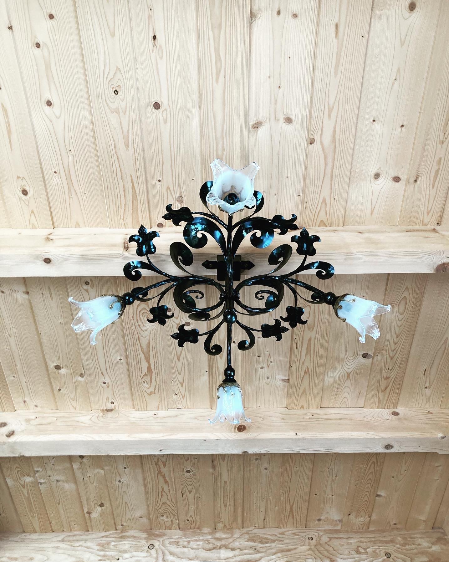 Kuta, metalowa konstrukcja z motywami roślinnymi i szklanymi, kwiatowymi kloszami, zamontowana na jasnym, drewnianym suficie z widocznymi belkami.