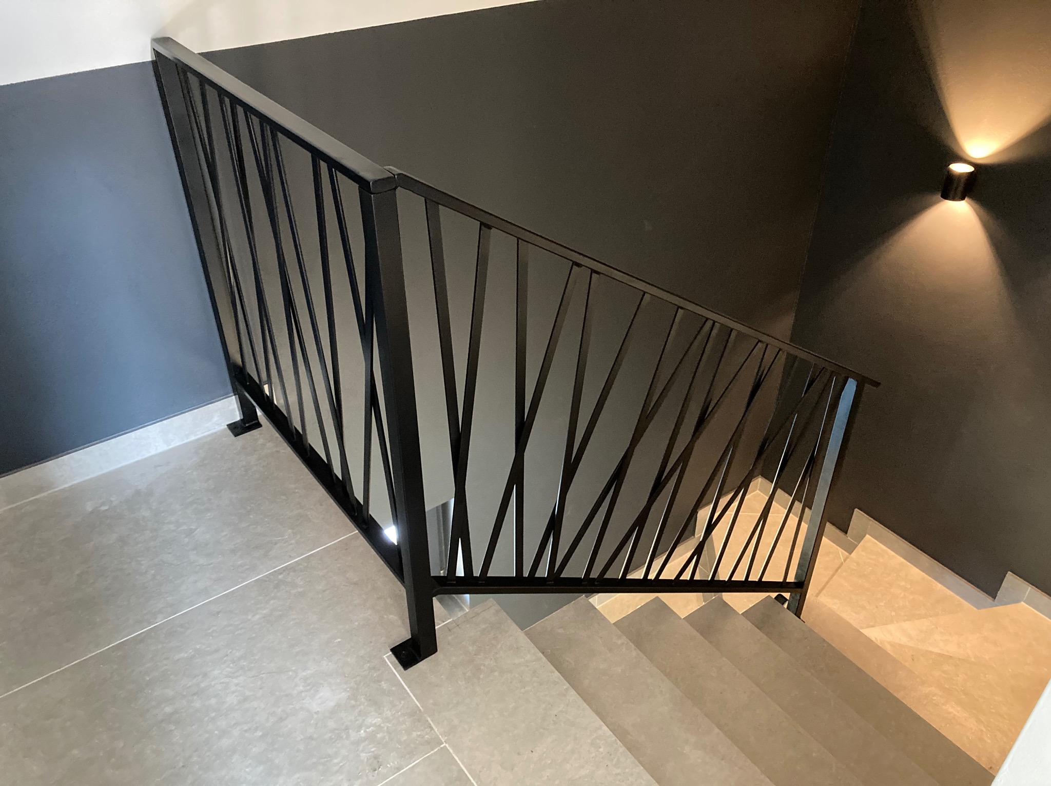 Nowoczesna, czarna poręcz schodowa z geometrycznym wypełnieniem z cienkich prętów, widok z góry na jasne, kafelkowe schody i ciemnoszarą ścianę z kinkietem.