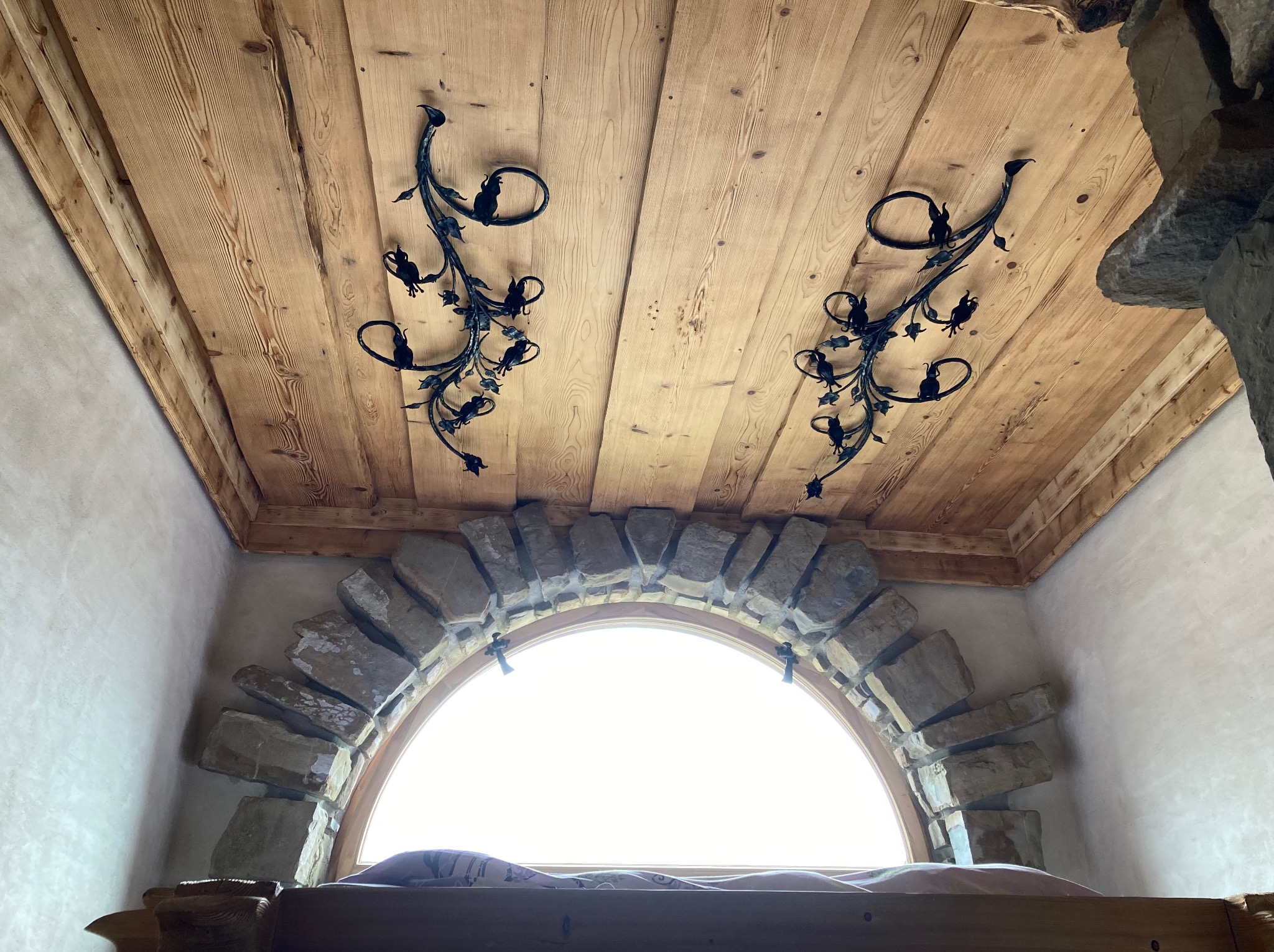 Drewniany sufit z dwoma kutymi, metalowymi dekoracjami w kształcie roślinnym nad łukowym oknem z kamienną ramą, widok z dołu.