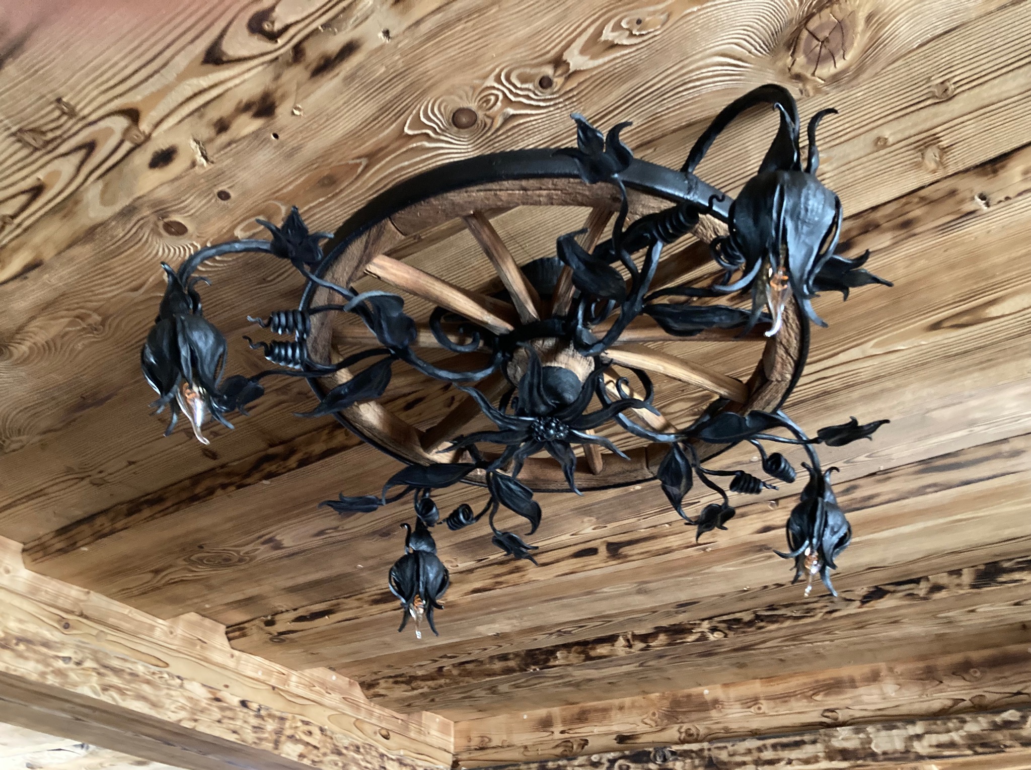 Ozdobny żyrandol z kutego metalu w kształcie kwiatów, zawieszony na drewnianym kole od wozu, zamontowany na drewnianym suficie z widocznymi słojami.