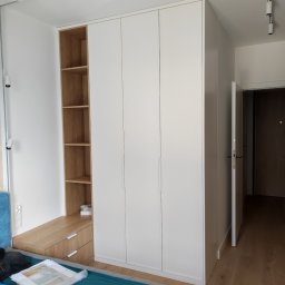 Karol Zysiak - Biała szafa z drewnianym regałem i szufladami w sypialni, widoczne oświetlenie sufitowe punktowe i fragment łóżka z niebieską narzutą.