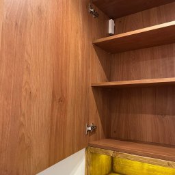 Karol Zysiak - Otwarta szafka z drewnianymi półkami i drzwiami o wyraźnym usłojeniu, dolna część podświetlona na żółto, widoczne zawiasy.