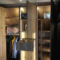 Karol Zysiak - Wnętrze przestronnej szafy z oświetleniem LED, otwartymi drzwiami po lewej, wysuwaną szufladą na środku, ubraniami na wieszakach, półkami z akcesoriami i sprzętami AGD, wykończonej w odcieniach...