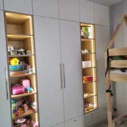 Biała szafa wnękowa z oświetlonymi półkami, wypełniona zabawkami i książkami dla dzieci, obok drewniane łóżko w kształcie domku.