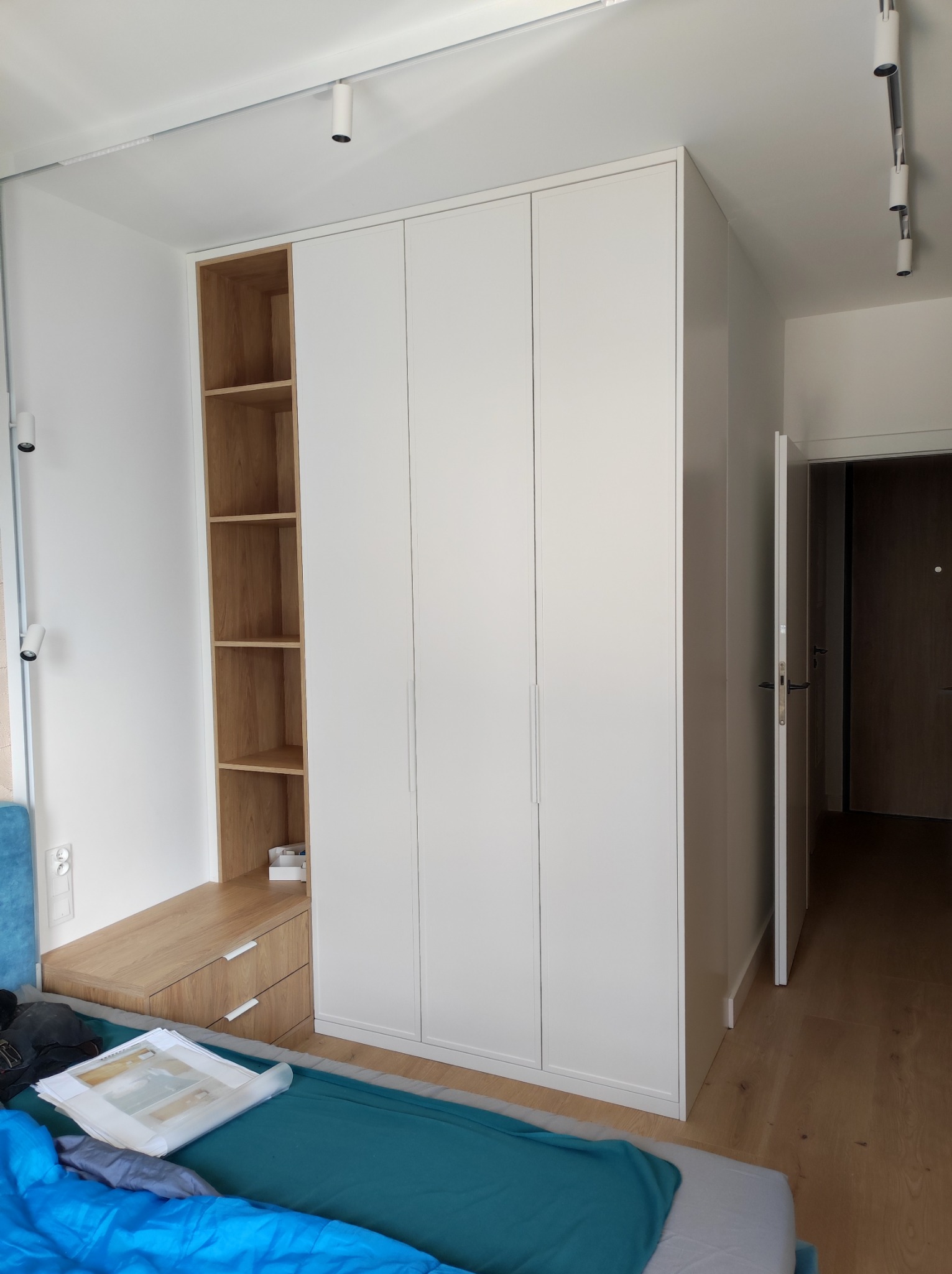 Biała szafa z drewnianym regałem i szufladami w sypialni, widoczne oświetlenie sufitowe punktowe i fragment łóżka z niebieską narzutą.