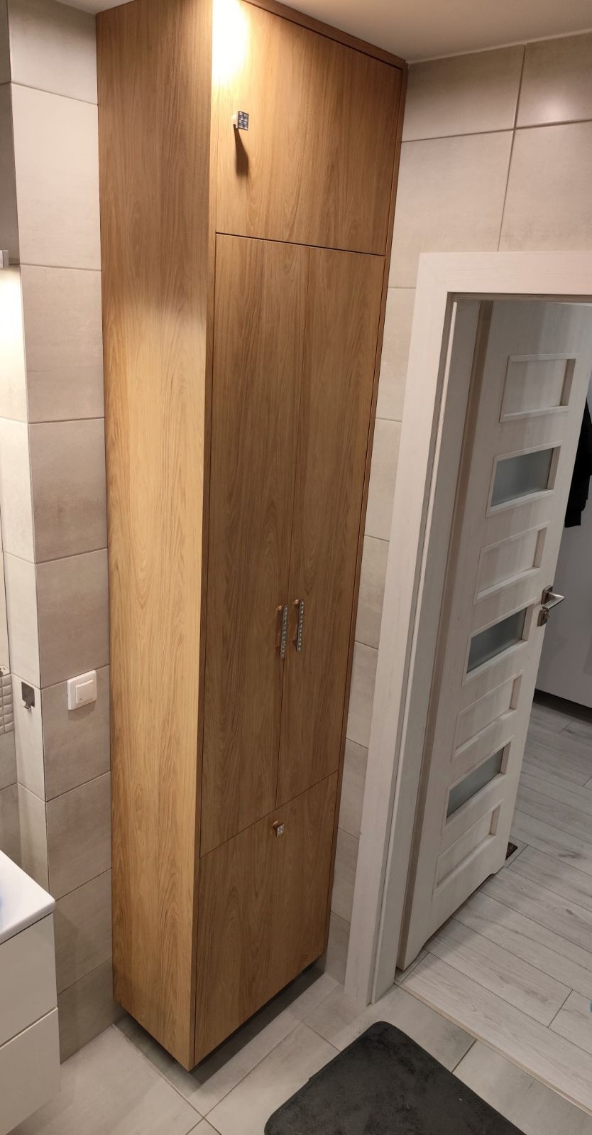 Wysoka, drewniana szafa na wymiar z dwiema parami uchwytów, zamontowana w łazience z jasnymi płytkami.