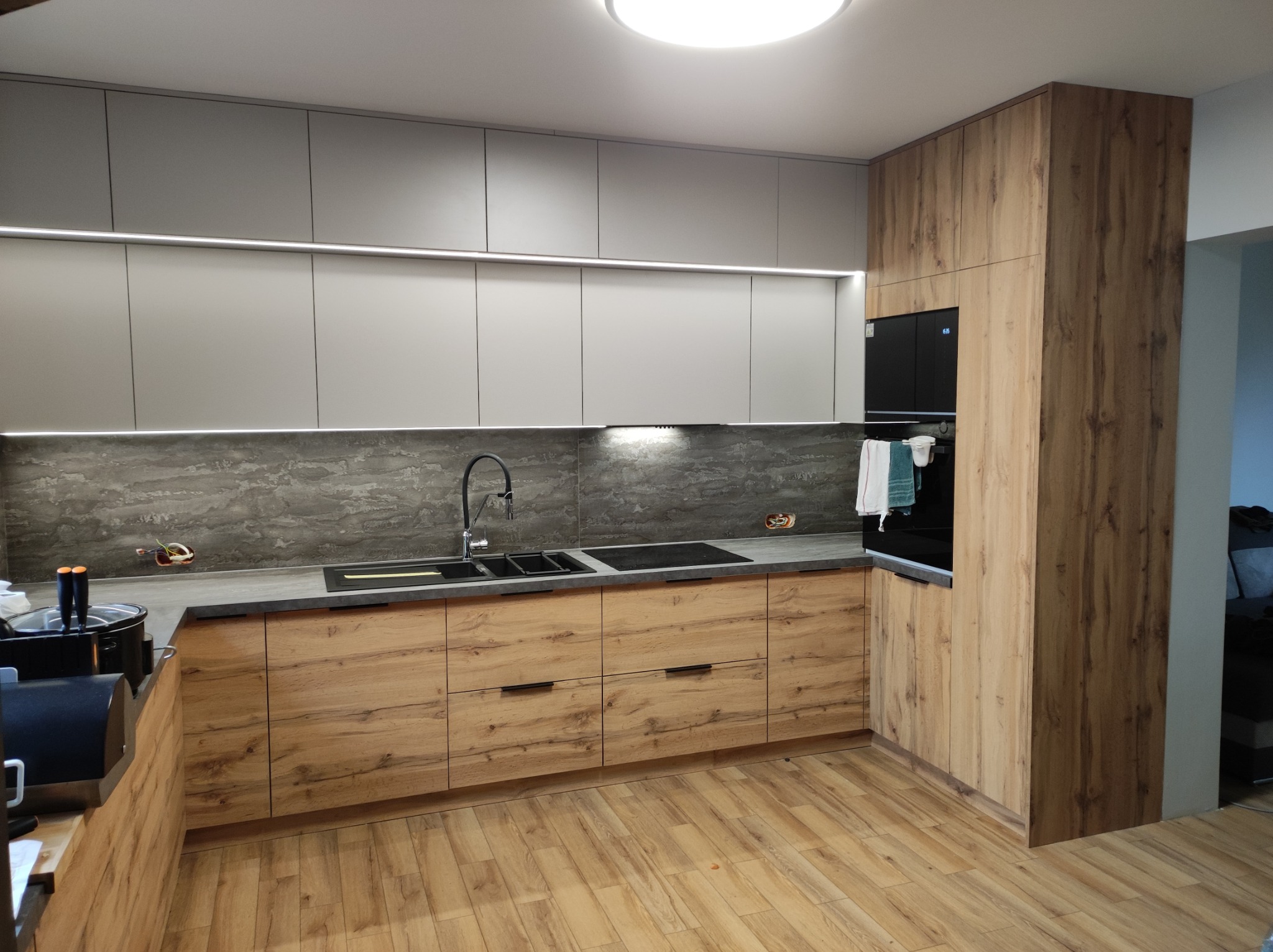 Nowoczesna kuchnia z drewnianymi frontami szafek dolnych i szarymi górnymi, czarny zlew i płyta indukcyjna wkomponowane w szary blat, oświetlenie LED pod szafkami wiszącymi, drewniana podłoga.