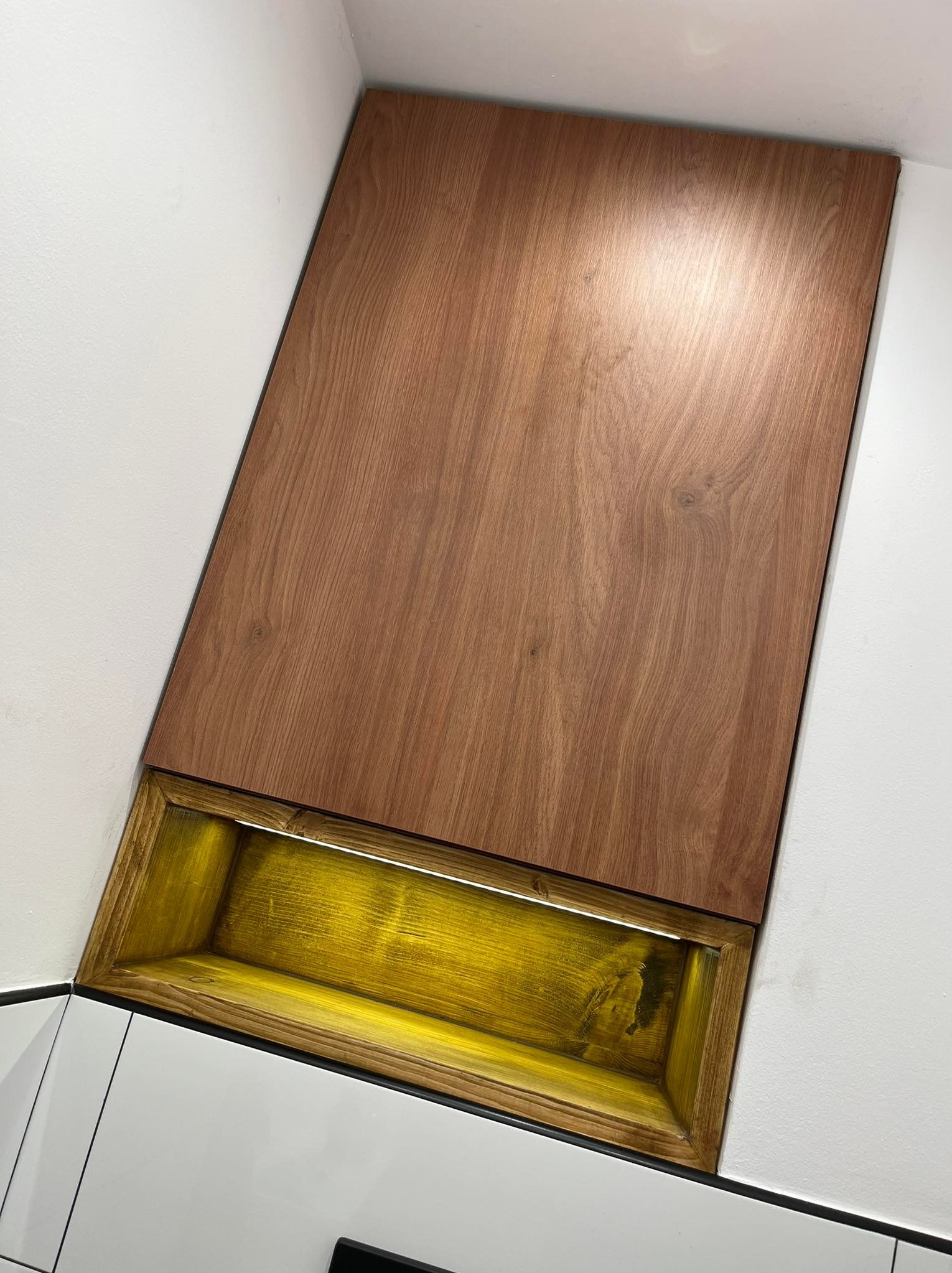 Fragment szafki kuchennej z drewnianym frontem i podświetlaną niszą w kolorze złota, zamontowanej pomiędzy białymi ścianami.