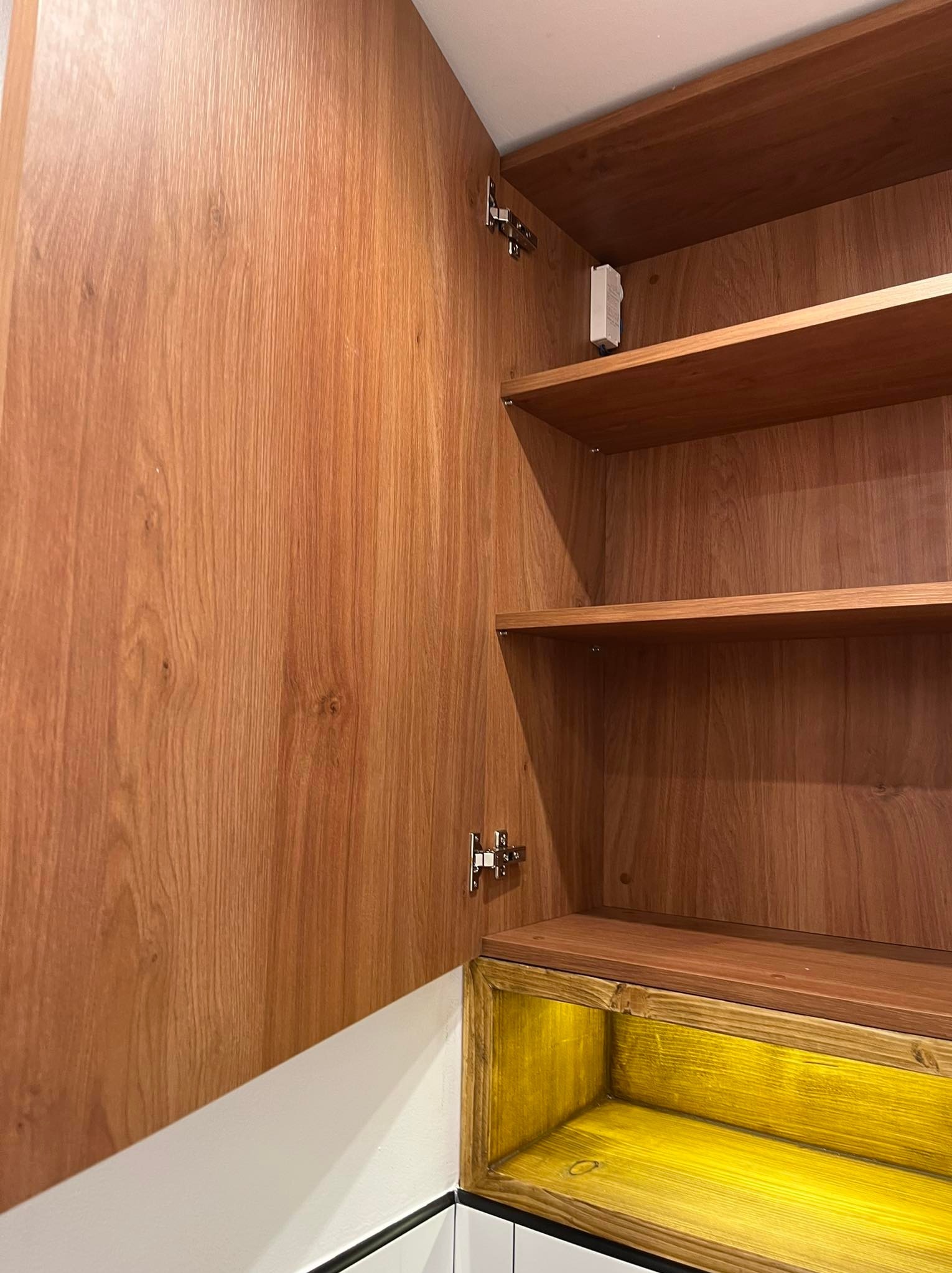 Otwarta szafka z drewnianymi półkami i drzwiami o wyraźnym usłojeniu, dolna część podświetlona na żółto, widoczne zawiasy.