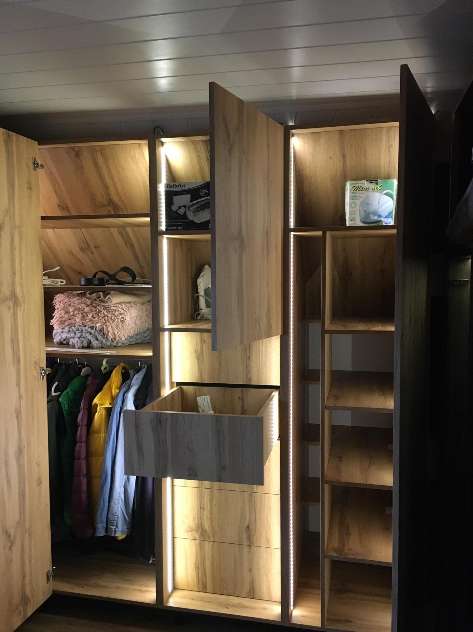Wnętrze przestronnej szafy z oświetleniem LED, otwartymi drzwiami po lewej, wysuwaną szufladą na środku, ubraniami na wieszakach, półkami z akcesoriami i sprzętami AGD, wykończonej w odcieniach...