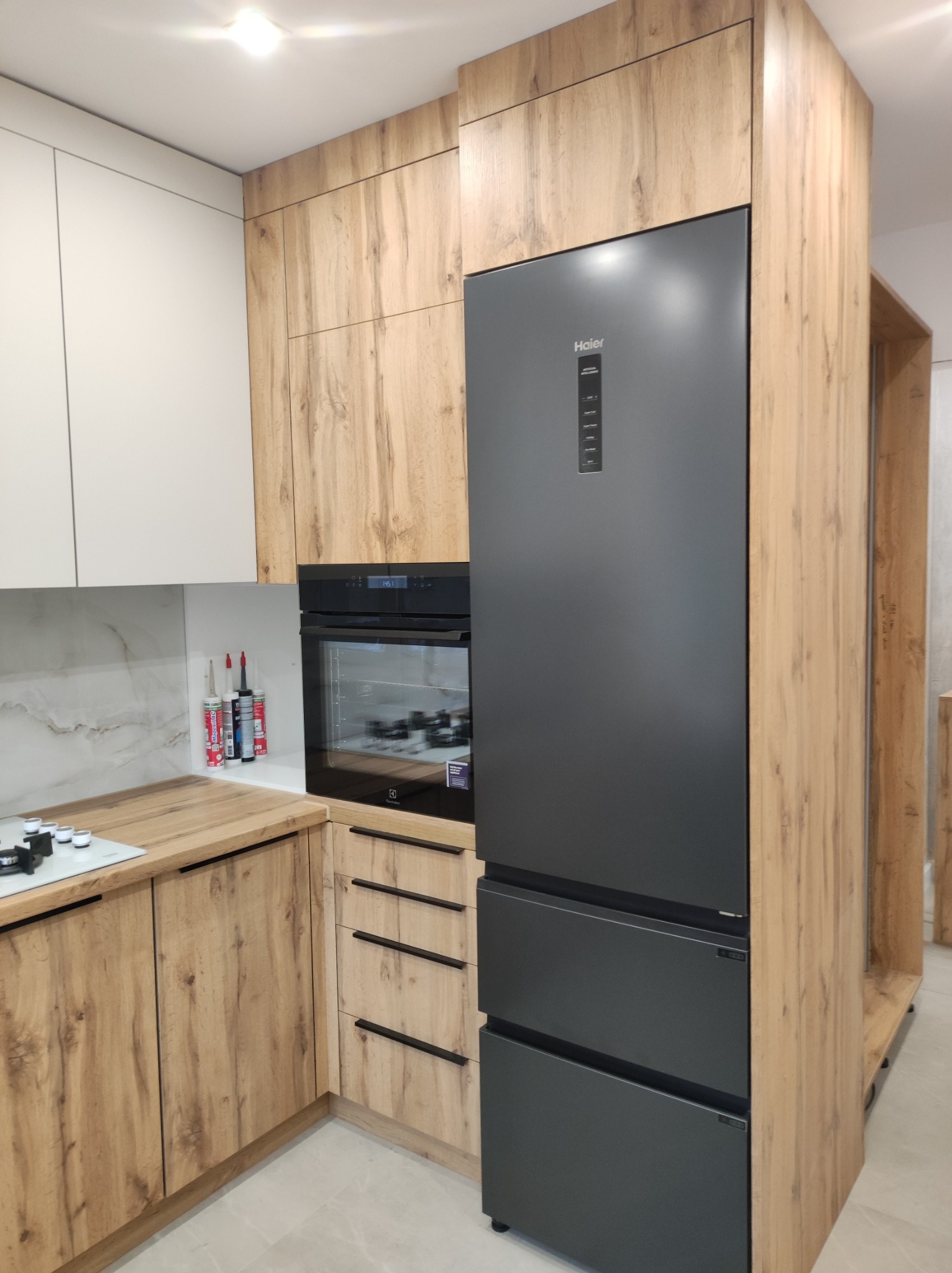 Nowoczesna zabudowa kuchenna z drewnopodobnymi frontami, grafitowa lodówka Haier, piekarnik Electrolux i białe szafki wiszące.