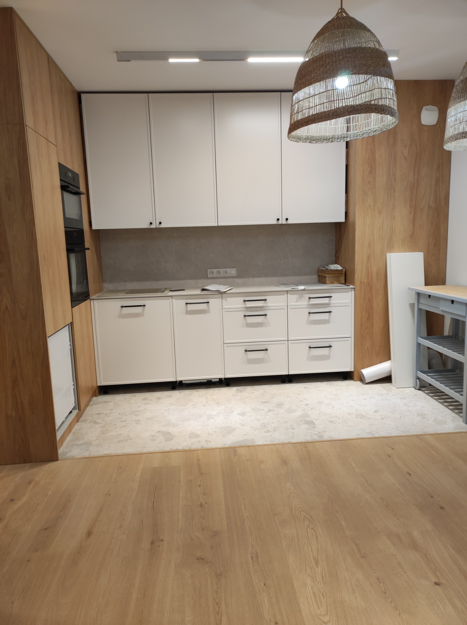 Nowoczesna kuchnia z białymi szafkami i drewnianymi panelami, oświetlona wiklinowymi lampami wiszącymi, z widocznym fragmentem podłogi w odcieniach drewna i marmurowej posadzki.