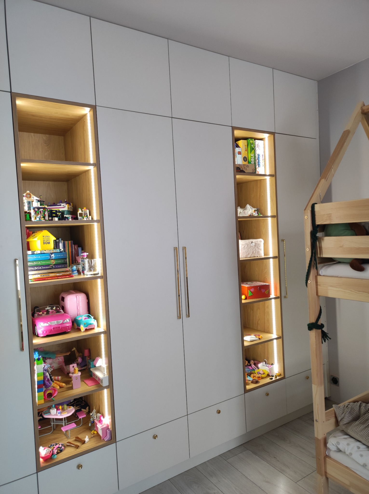 Biała szafa wnękowa z oświetlonymi półkami, wypełniona zabawkami i książkami dla dzieci, obok drewniane łóżko w kształcie domku.