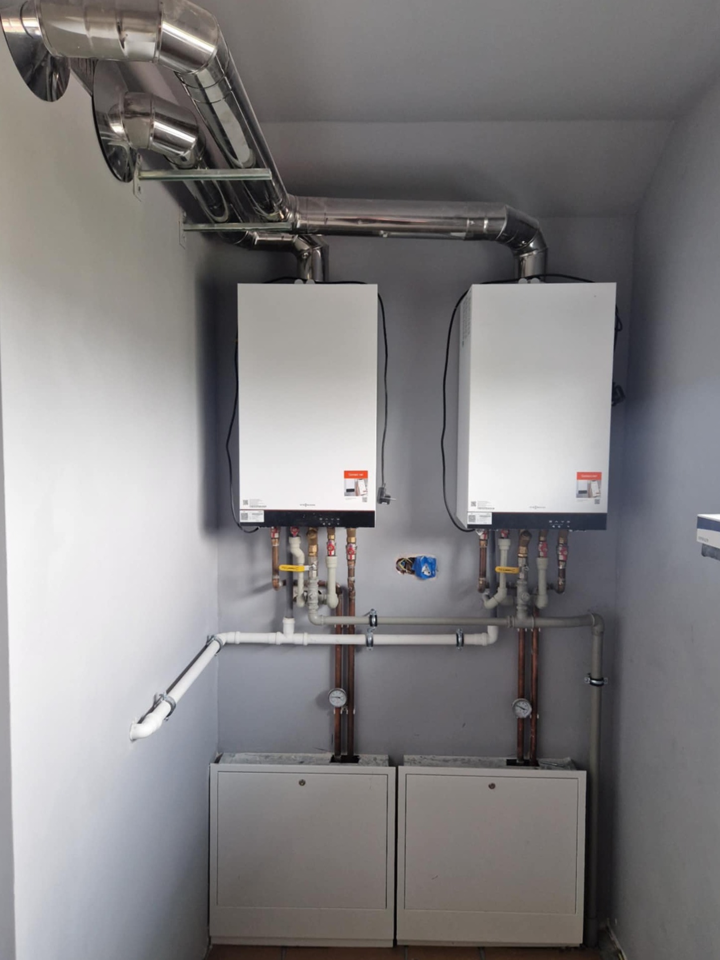 Dwa białe piece gazowe zamontowane na szarej ścianie, z widocznymi rurami miedzianymi i białymi, oraz srebrną rurą odprowadzającą spaliny pod sufitem.