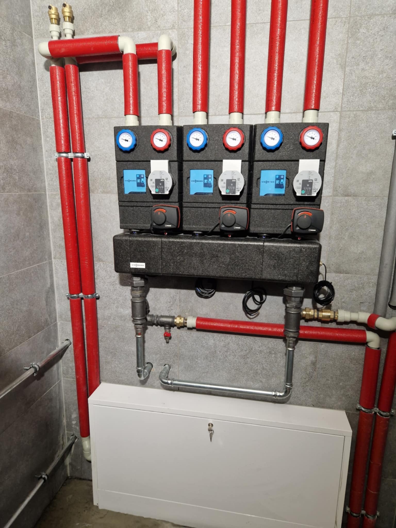 Rozdzielacz ogrzewania podłogowego z czterema obiegami, każdy wyposażony w pompę, zawory termostatyczne i wskaźniki temperatury, zamontowany na szarej ścianie z rurami w izolacji koloru czerwonego.