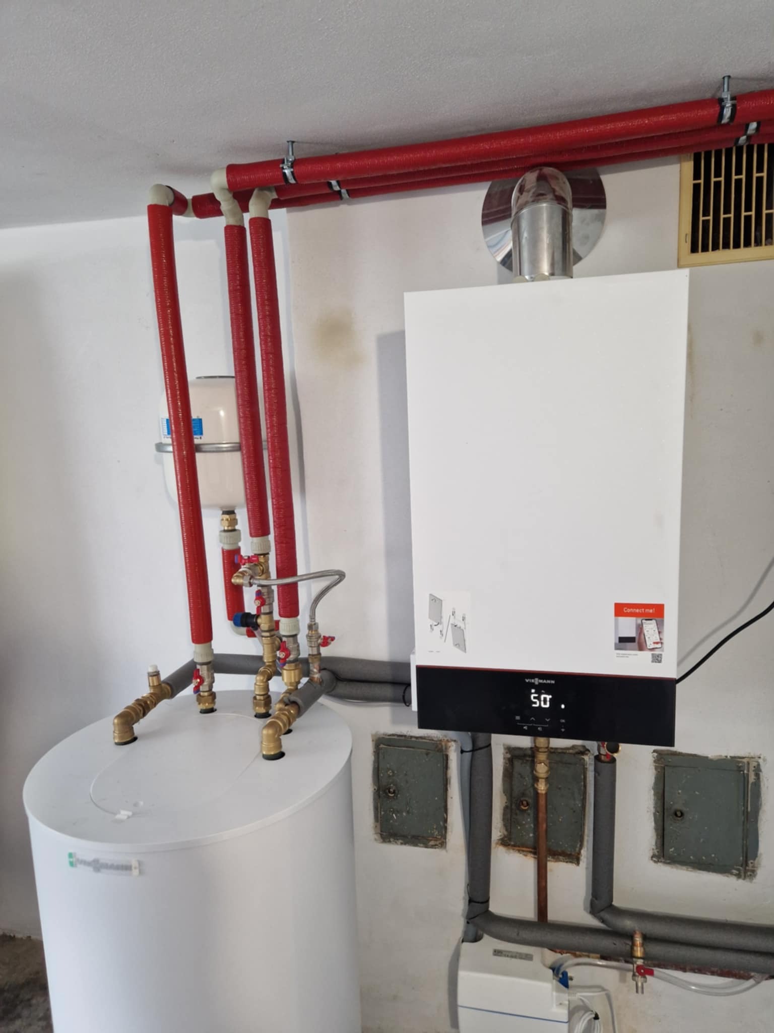 Biały piec gazowy Viessmann z podłączonymi rurami miedzianymi i czerwonymi rurami izolowanymi, zamontowany na białej ścianie obok białego zasobnika ciepłej wody z widocznymi zaworami.