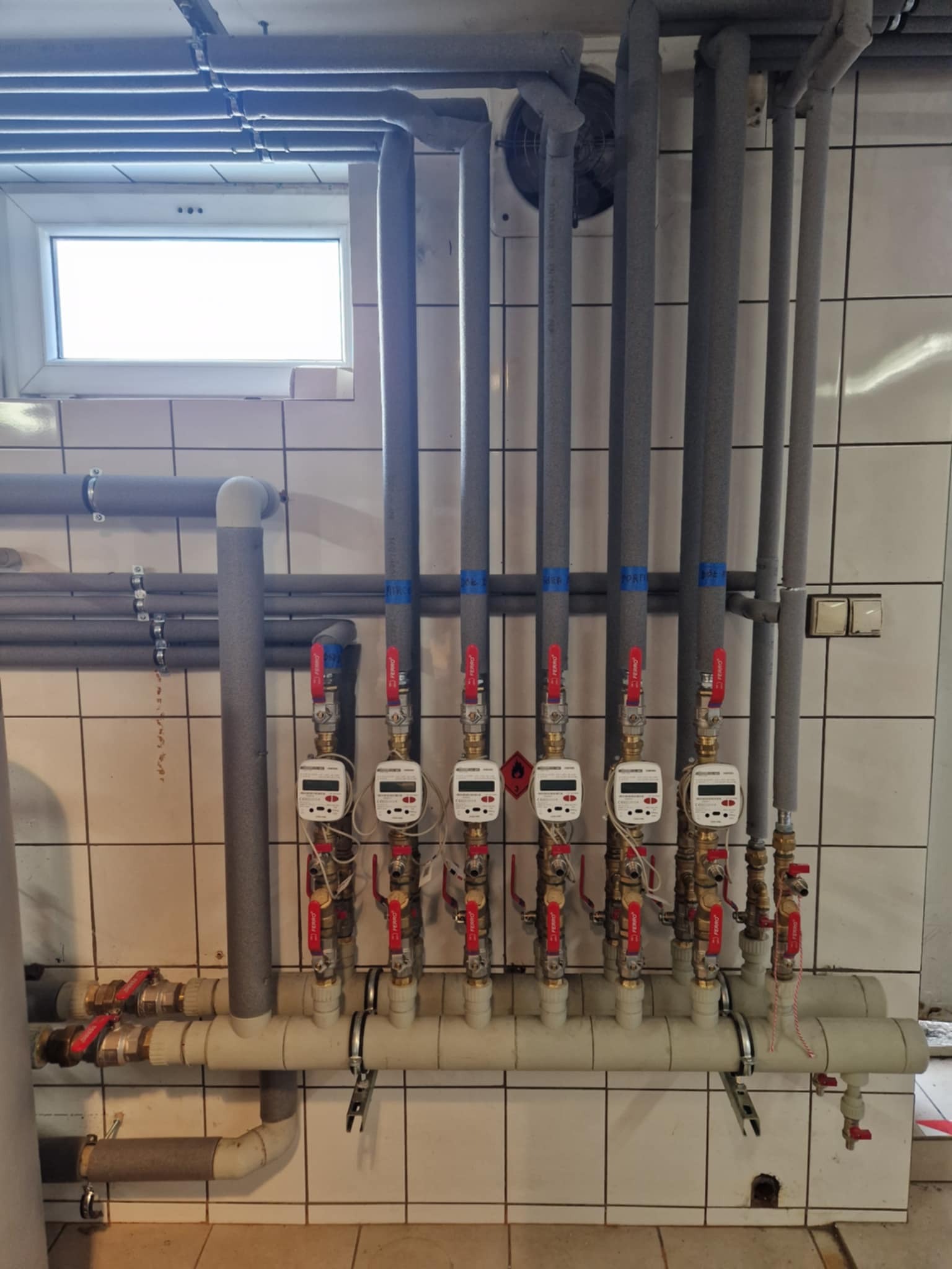 Szereg pionowych rur w izolacji, z zamontowanymi licznikami ciepła z czerwonymi zaworami, na tle białych płytek ceramicznych.