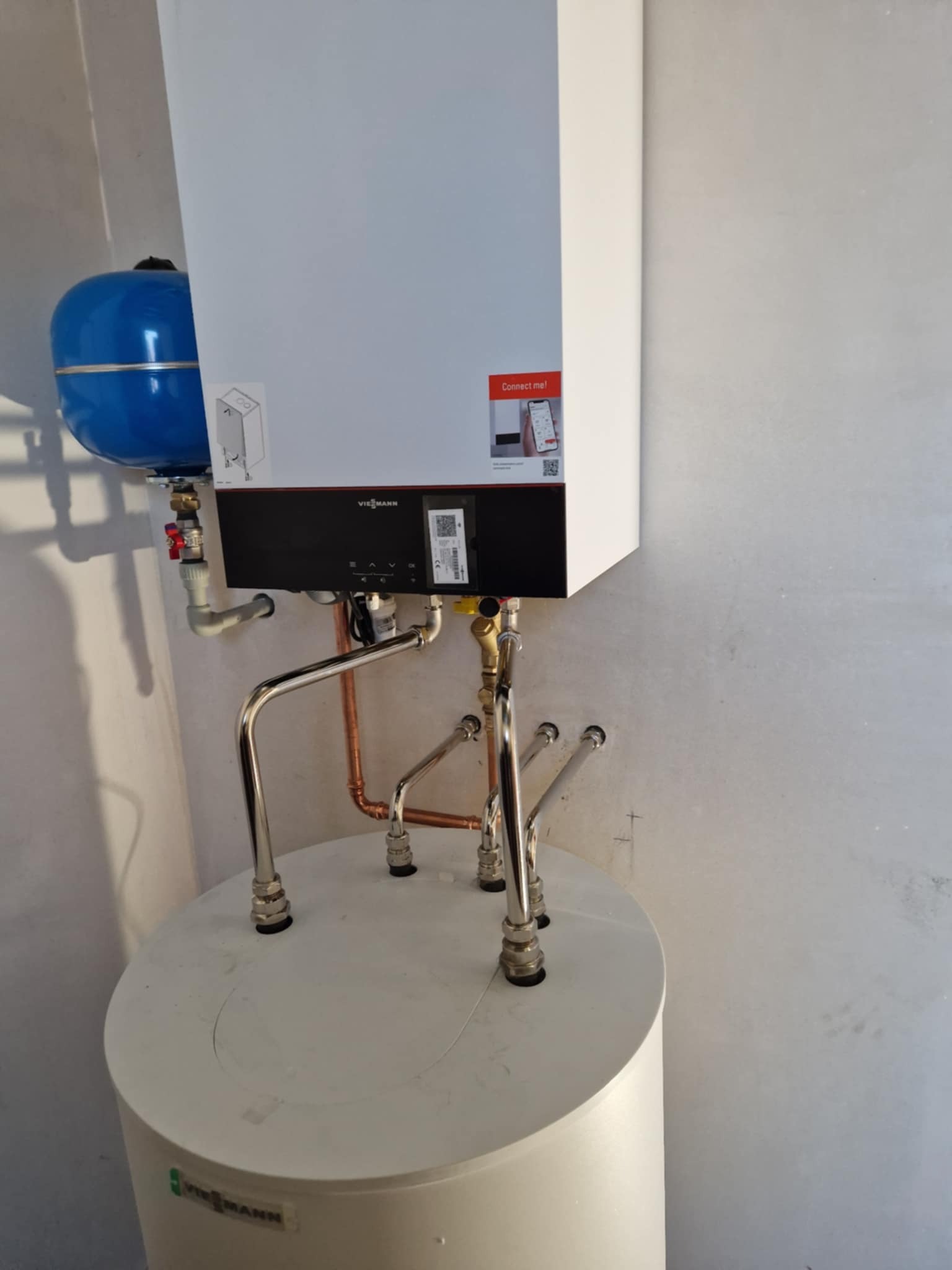 Nowoczesny piec gazowy marki Viessmann, zamontowany na białym zasobniku, z widocznymi miedzianymi i chromowanymi rurami przyłączeniowymi oraz niebieskim naczyniem przeponowym.