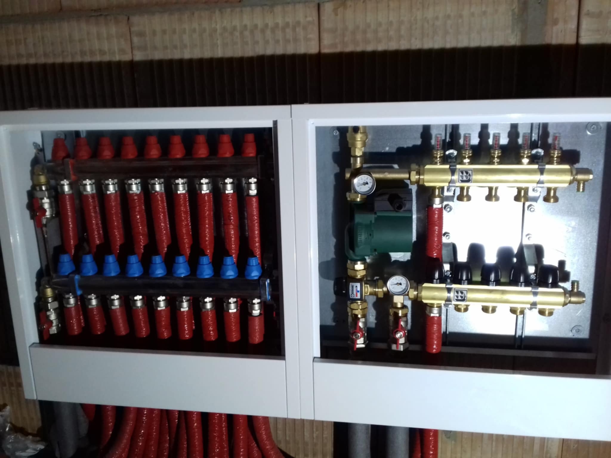 Rozdzielacz ogrzewania podłogowego w białej obudowie, z widocznymi czerwonymi rurami i elementami regulacyjnymi z manometrami, zainstalowany na tle ściany z pustaków.