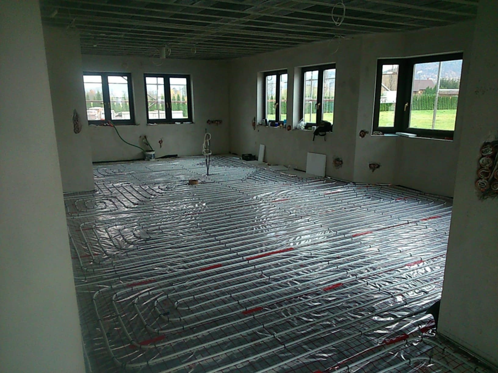 Wnętrze w trakcie instalacji ogrzewania podłogowego, widoczne rury grzewcze rozłożone na folii izolacyjnej, surowy stan wykończenia ścian i sufitu, okna z widokiem na zieleń.