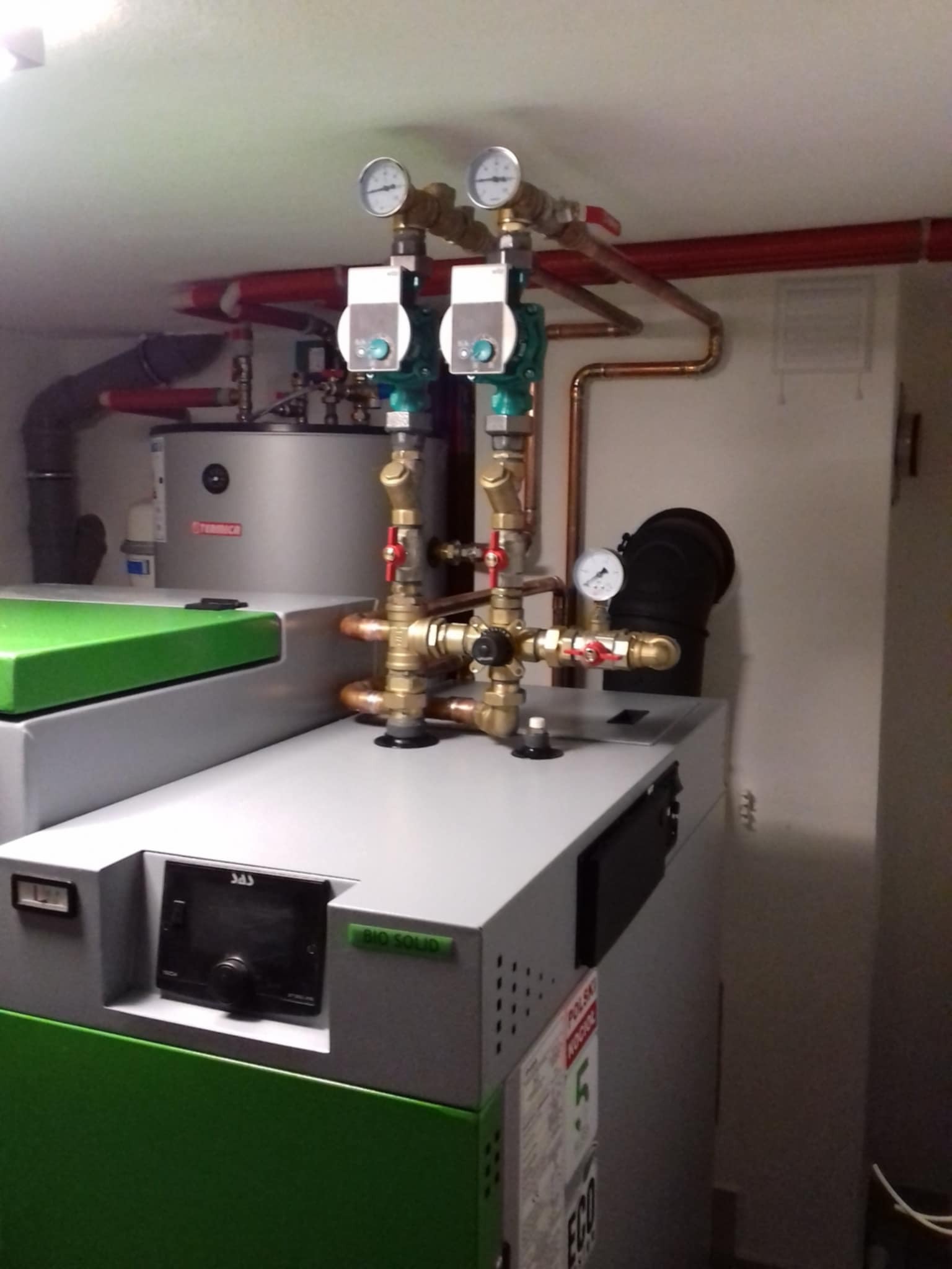 Nowoczesny piec na paliwo stałe z zielonym akcentem i widocznymi elementami instalacji grzewczej, w tym zaworami i manometrami, zainstalowany w pomieszczeniu technicznym.