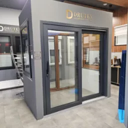 Ekspozycja w salonie: przesuwne okno tarasowe z ciemną ramą, widoczne logo producenta Drutex, w tle inne okna i rolety.